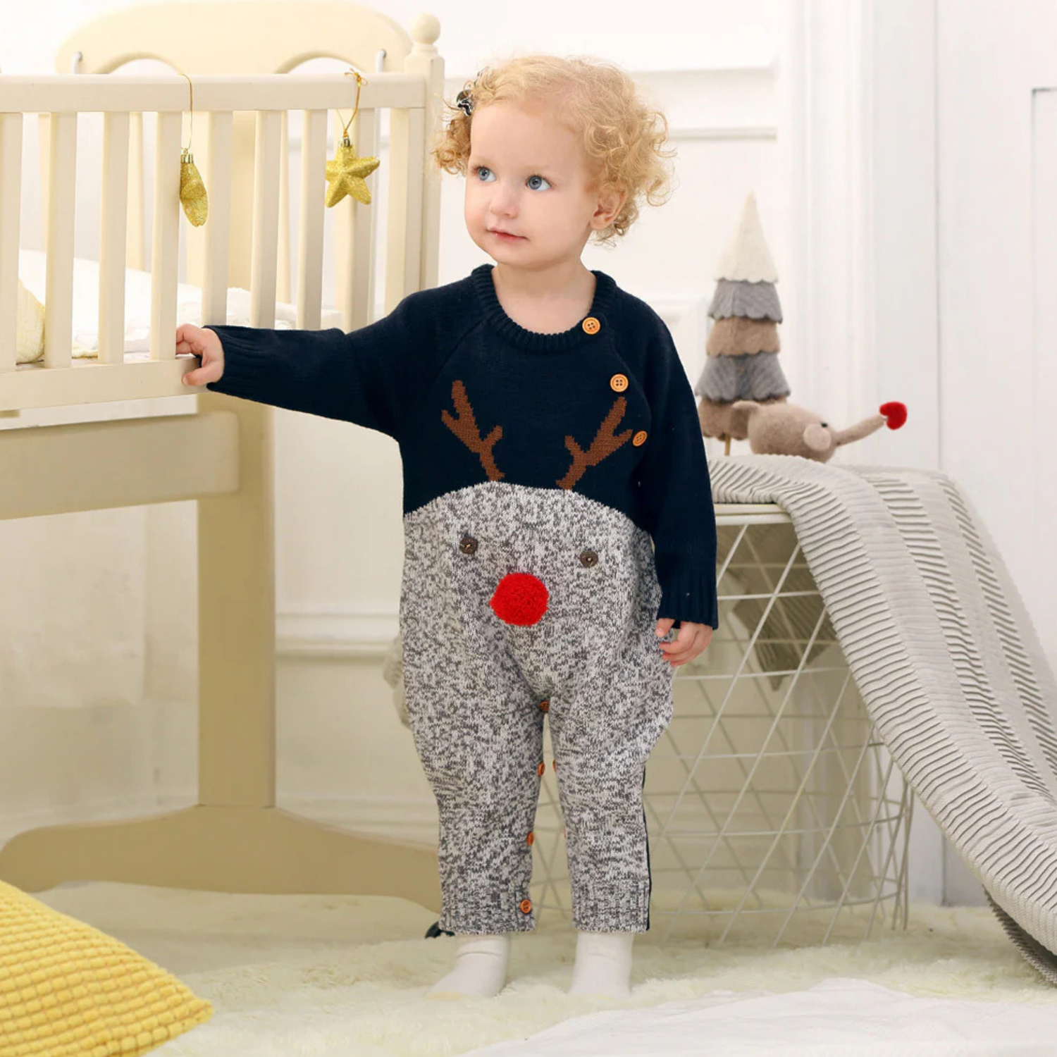 Bébé portant une combinaison de Noël bleue et grise avec motif renne et pompon rouge, debout près d'un lit décoré.