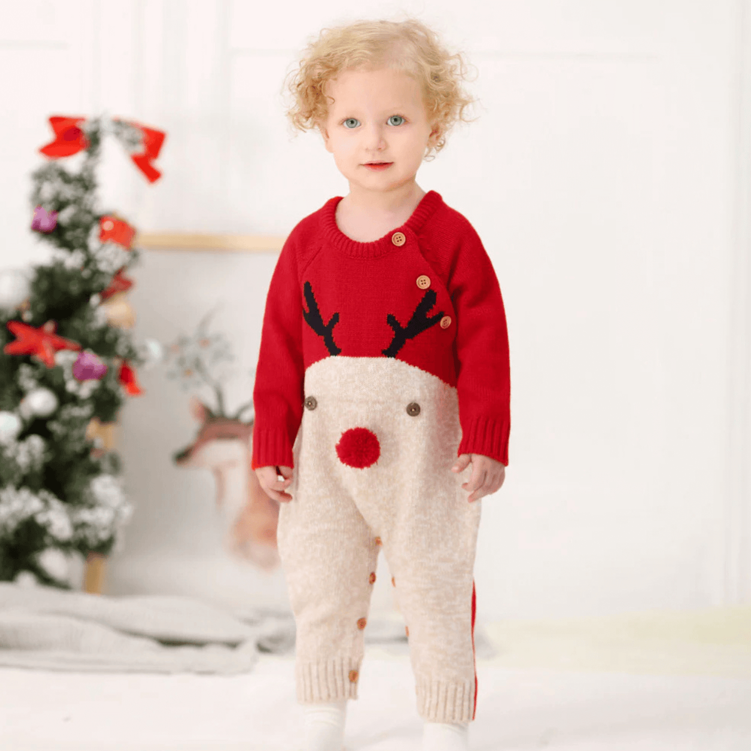 Bébé en combinaison rouge avec motif renne et pompon rouge, debout près d'un sapin de Noël décoré